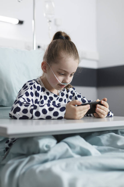 Sick young girl watching internet funny videos on smart mobile device while resting on clinic bed - Fotó, kép