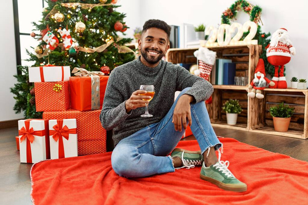 Junger afrikanisch-amerikanischer Mann lächelt glücklich bei der Weihnachtsfeier und trinkt Champagner zu Hause. - Foto, Bild