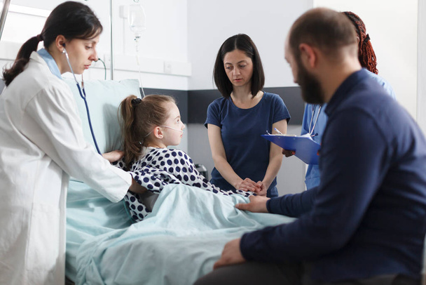 Pediatra doradztwo chorej dziewczynki za pomocą stetoskopu, podczas gdy zmartwionych rodziców czeka obok niej. - Zdjęcie, obraz
