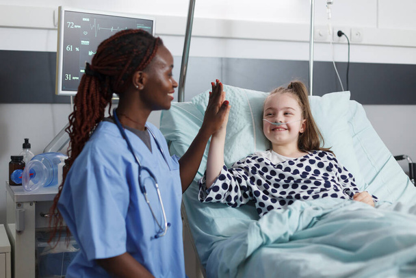 Healthcare clinic nurse highfive ill little girl while in patient recovery ward room. - Zdjęcie, obraz