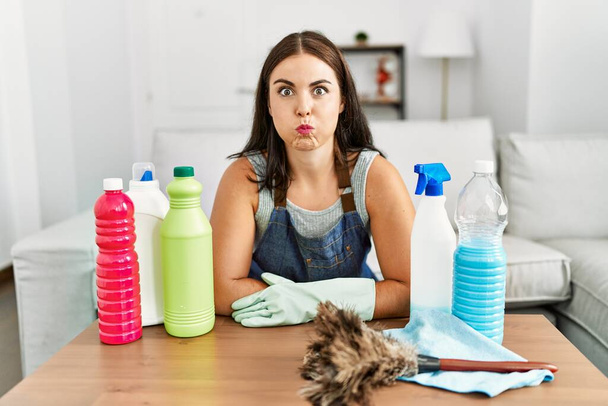 Young brunette woman wearing cleaner apron and gloves cleaning at home puffing cheeks with funny face. mouth inflated with air, crazy expression.  - Photo, Image