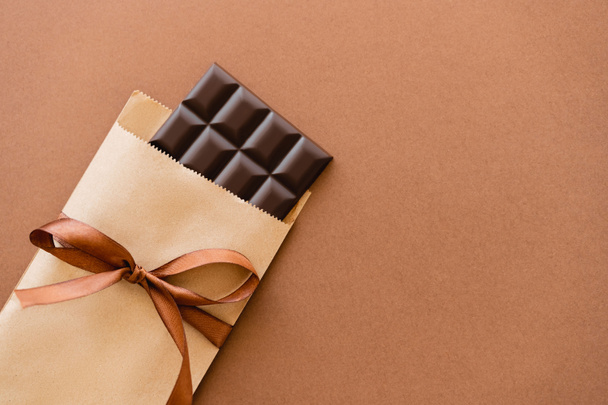 Top view of dark chocolate bar in craft package with ribbon on brown background  - Photo, Image
