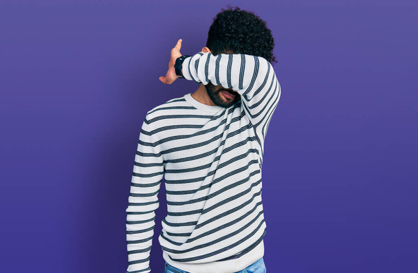 Young arab man with beard wearing casual striped sweater covering eyes with arm, looking serious and sad. sightless, hiding and rejection concept  - Photo, Image