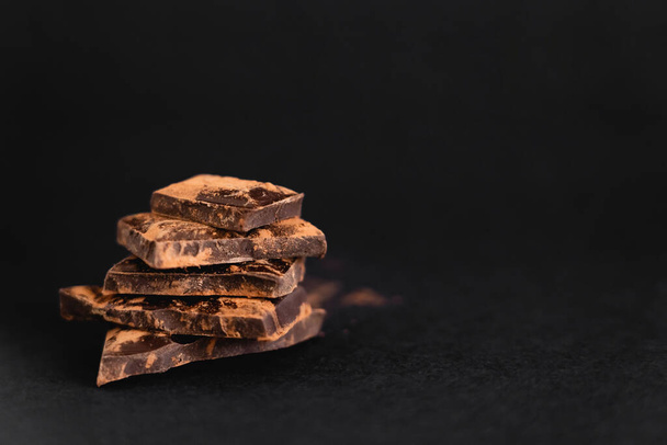 Close-up zicht op stukjes pure chocolade met droge cacao op zwarte ondergrond - Foto, afbeelding