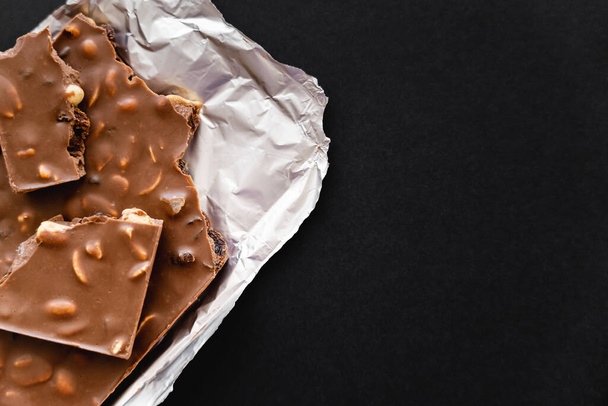 Top view of natural milk chocolate with nuts on foil isolated on black  - Photo, image