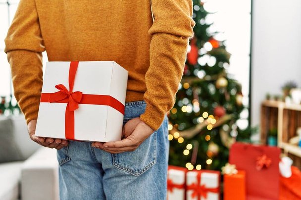 Hombre sosteniendo el regalo en su espalda de pie junto al árbol de Navidad en casa. - Foto, Imagen