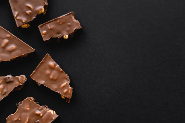 Top view of pieces of milk chocolate with nuts on black surface with copy space - Photo, Image