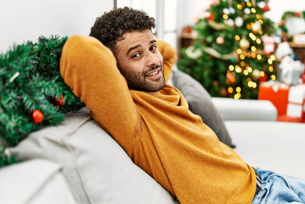 Young arab man relaxed with hands on head sitting on the sofa by christmas tree at home. - Foto, Bild