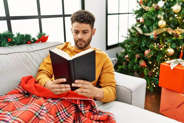Young arab man reading book lying on the sofa by christmas tree at home. - Foto, afbeelding