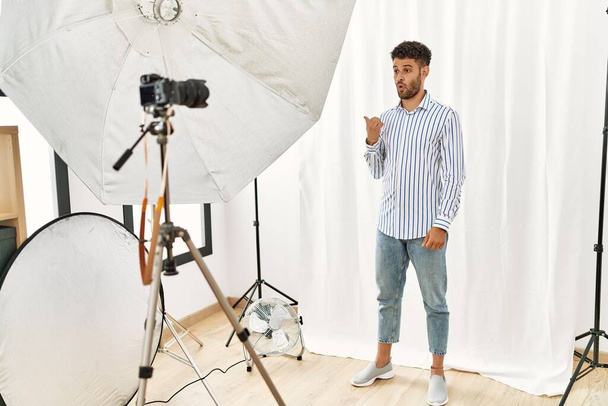Hombre joven árabe haciéndose pasar por modelo en el estudio de fotografía sorprendido señalando con el dedo de la mano hacia un lado, boca abierta expresión asombrada.  - Foto, Imagen