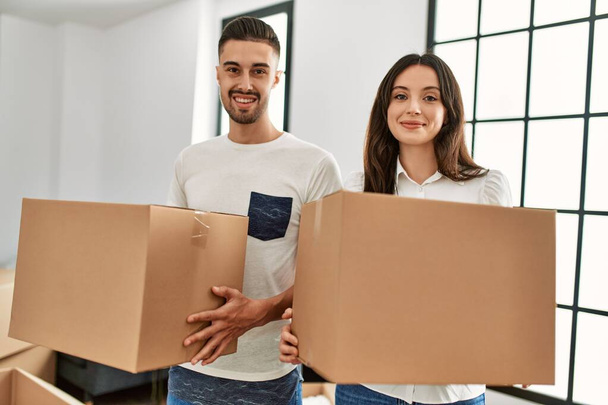 Jovem casal hispânico sorrindo feliz segurando caixas de papelão se movendo em nova casa. - Foto, Imagem