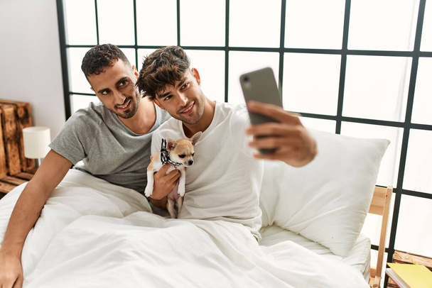 Two hispanic men couple make selfie by the smartphone sitting on bed with chihuahua at bedroom - Fotografie, Obrázek