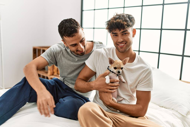 Dos hombres hispanos se abrazan sentados en la cama con chihuahua en el dormitorio - Foto, Imagen