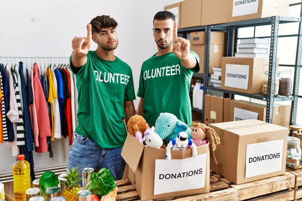 Young gay couple wearing volunteer t shirt at donations stand pointing with finger up and angry expression, showing no gesture  - Photo, Image