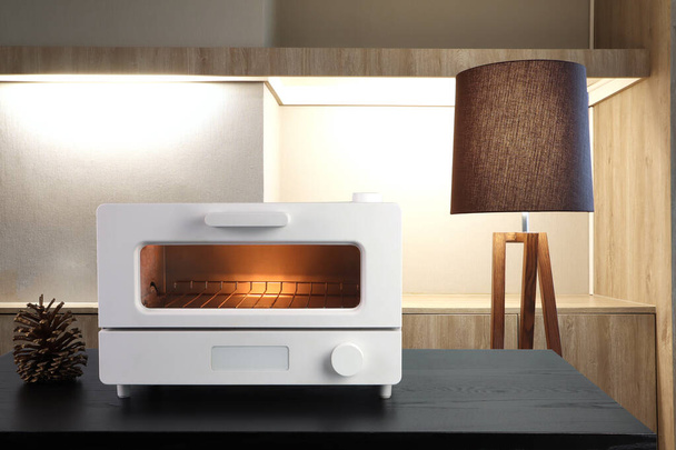 white modern design toaster oven is on the black wooden table for making bread with background of nice luxury kitchen room with beautiful lamp during breakfast in the morning - Foto, afbeelding