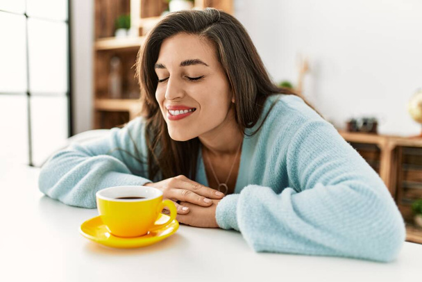 Молодая женщина нюхает кофе сидя дома на столе - Фото, изображение
