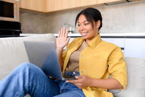 Multiracial woman wearing domestic clothes sitting in the living room, relaxing and talking to her friend or family, waving at the laptop screen - Foto, Imagem