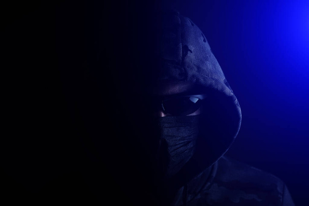 Stylish man in dark hoodie, black glasses and mask dark background in blue light, hacker, attacker and bandit - Foto, Bild