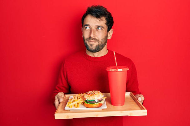 Hombre guapo con barba comiendo una sabrosa hamburguesa clásica con papas fritas y refrescos sonriendo mirando a un lado y mirando hacia otro pensando.  - Foto, imagen