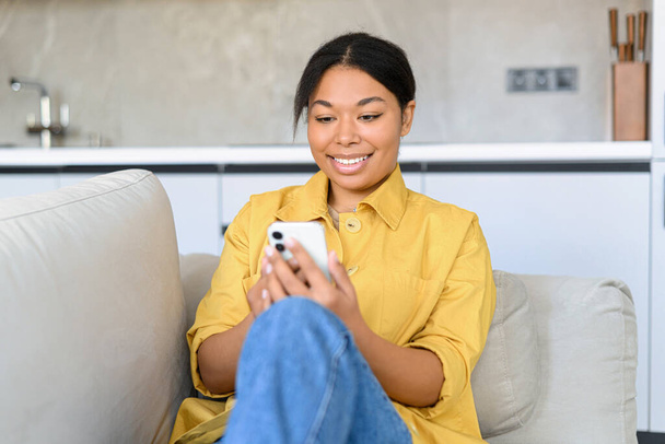 Happy multiracial woman sitting on the couch and spending time online with her smartphone, texting messages, scrolling news feed, sharing post in social networks - Фото, изображение
