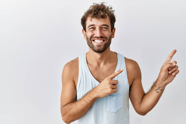 Jonge knappe man staat over geïsoleerde achtergrond glimlachend en kijkend naar de camera wijzen met twee handen en vingers naar de zijkant.  - Foto, afbeelding