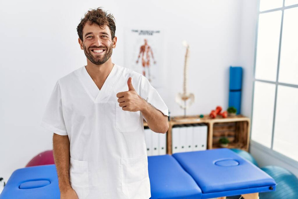 Young handsome physiotherapist man working at pain recovery clinic doing happy thumbs up gesture with hand. approving expression looking at the camera showing success.  - Photo, image