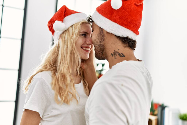 Casal jovem sorrindo feliz em atitude romântica usando chapéu de Natal em casa. - Foto, Imagem