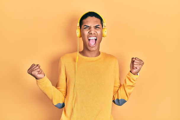 Young african american guy listening to music using headphones crazy and mad shouting and yelling with aggressive expression and arms raised. frustration concept.  - Foto, afbeelding