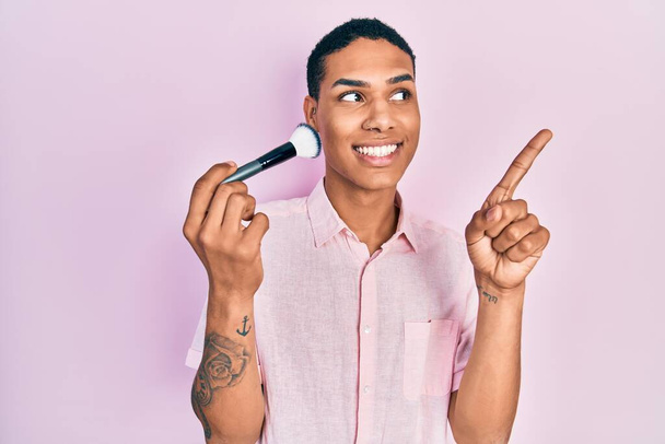 Young african american guy holding makeup brush smiling happy pointing with hand and finger to the side  - Foto, afbeelding