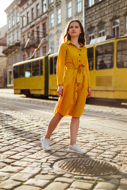 Stijlvol schattig meisje in de zomer shirt jurk en oude tram. Zomer vrouw portret in gele outfit op stad achtergrond - Foto, afbeelding