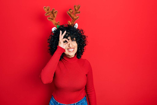 Young middle east woman wearing cute christmas reindeer horns doing ok gesture with hand smiling, eye looking through fingers with happy face.  - Foto, immagini