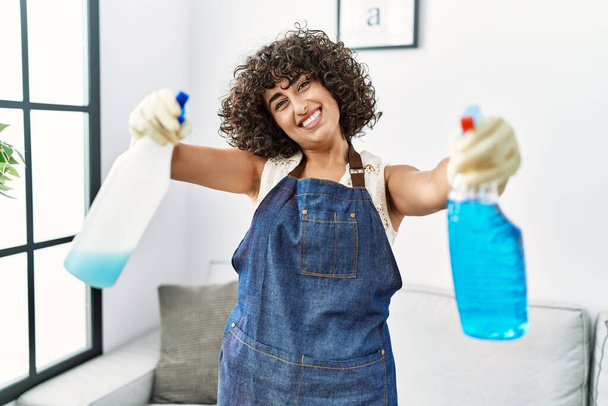 Joven mujer de Oriente Medio sonriendo confiado sosteniendo pulverizador de limpieza en casa - Foto, imagen