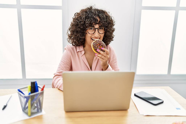 Молода близькосхідна бізнес-леді їсть пончик, що працює в офісі
. - Фото, зображення