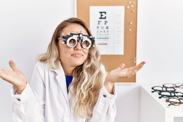 Young beautiful optician woman wearing optometry glasses at the clinic clueless and confused expression with arms and hands raised. doubt concept.  - Photo, Image