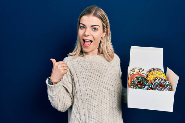 Красива біла жінка тримає коробку смачних барвистих пончиків, спрямованих на великий палець в сторону посміхаючись з відкритим ротом
  - Фото, зображення