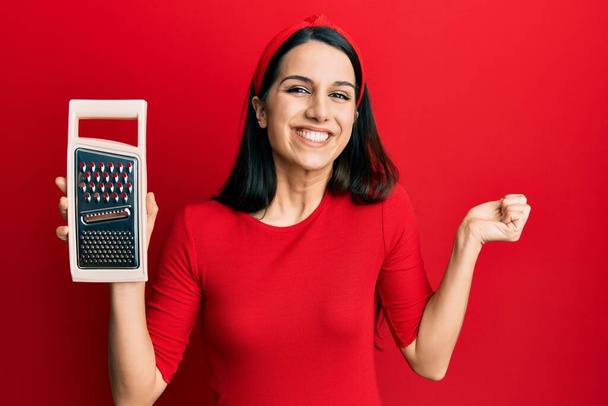 Young hispanic woman holding grater screaming proud, celebrating victory and success very excited with raised arm  - Fotó, kép