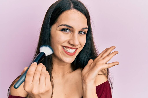 Young hispanic woman holding makeup brush and blush celebrating achievement with happy smile and winner expression with raised hand  - Foto, Imagen
