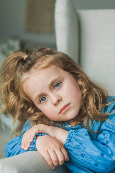 A little cute blue-eyed girl with curly long hair sits in a cozy armchair at home. Cozy, bright house. Happy child is resting. Home. - Fotografie, Obrázek