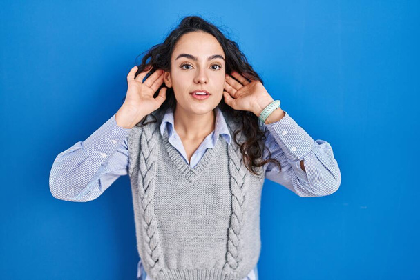 Young brunette woman standing over blue background trying to hear both hands on ear gesture, curious for gossip. hearing problem, deaf  - Zdjęcie, obraz