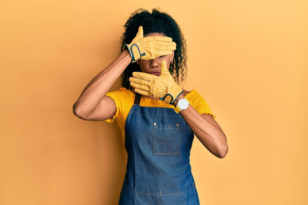 Femme afro-américaine du Moyen Âge portant tablier professionnel couvrant les yeux et la bouche avec les mains, surpris et choqué. cacher l'émotion  - Photo, image