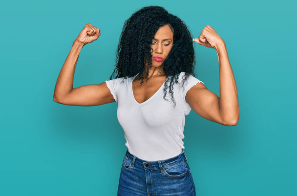 Mujer afroamericana de mediana edad que usa una camiseta blanca casual que muestra los músculos de los brazos sonriendo orgullosa. concepto de fitness.  - Foto, Imagen
