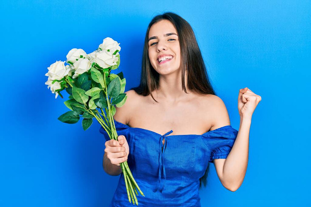 Young brunette teenager holding bouquet of white flowers screaming proud, celebrating victory and success very excited with raised arm  - Photo, Image