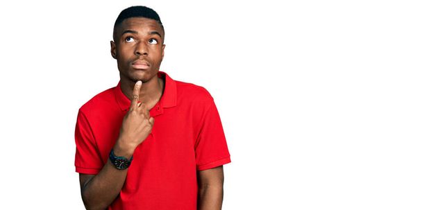 Genç Afro-Amerikalı bir adam günlük kırmızı tişört giyiyordu. Düşünceleri kuşkuya yoğunlaşmıştı. Parmakları çenesinde ve yukarı bakıyordu.  - Fotoğraf, Görsel