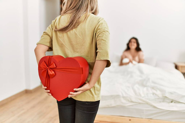 Young woman surprising her girlfriend with valentine gift at bedroom. - Фото, изображение