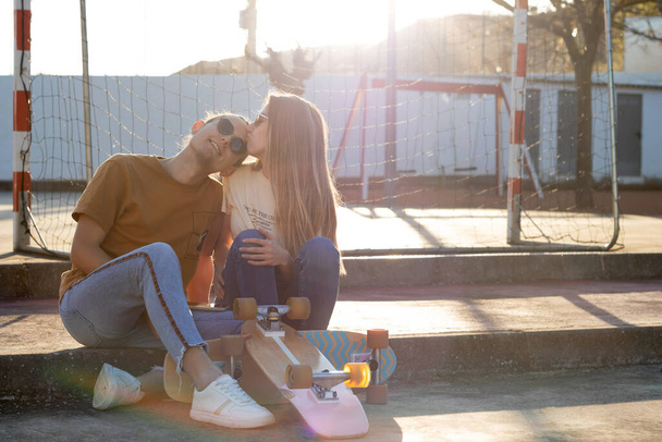 Cute lovely heterosexual couple sitting kissing on forehead with sun behind them after going on long board - Foto, Imagem