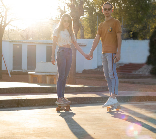 Πανέμορφο νεαρό καυκάσιο ζευγάρι να κάνει πατινάζ μαζί στις σανίδες των καταδρομικών τους κρατώντας τα χέρια το ηλιοβασίλεμα - Φωτογραφία, εικόνα