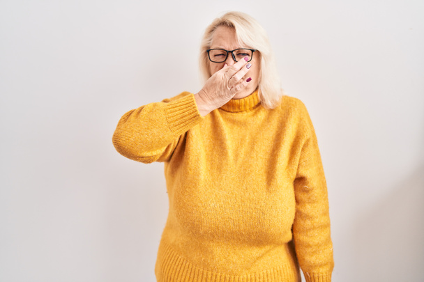 Mujer caucásica de mediana edad con gafas de pie sobre el fondo oliendo algo apestoso y asqueroso, olor intolerable, conteniendo la respiración con los dedos en la nariz. mal olor  - Foto, imagen