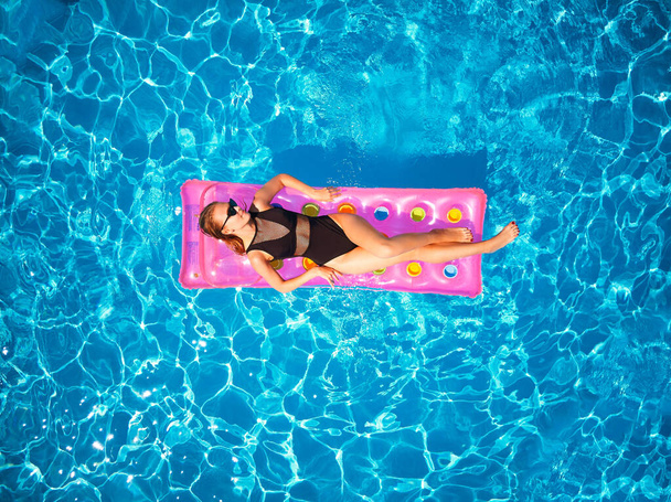 Fit pretty girl in bikini chilling on inflatable pink mattress in swimming pool. Slim hot woman in swimwear tanning. Female relaxing on float in blue water at luxury resort. Aerial, view from above. - Photo, Image