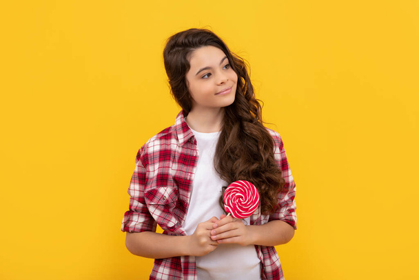 smiling kid with long curly hair hold lollipop caramel candy on yellow background, sweet tooth. - Valokuva, kuva