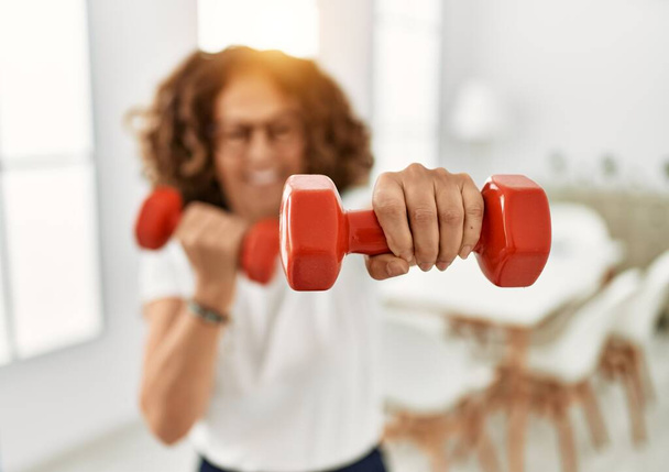 Mujer hispana de mediana edad sonriendo entrenando confiadamente usando pesas en casa - Foto, imagen
